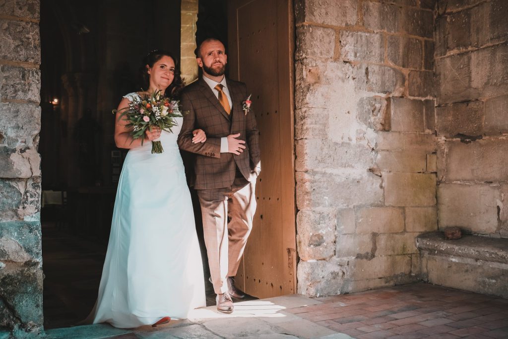 entrée des mariés sortant d’une église en pierre, la mariée au bras de son marié devant la grande porte en bois