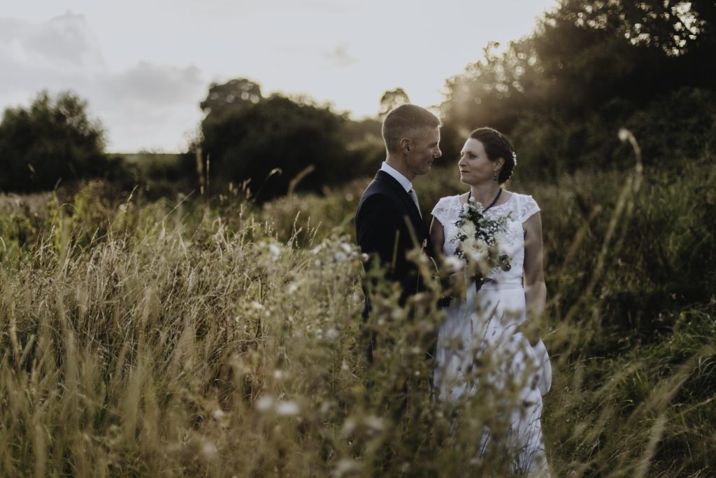 mariés debout dans une prairie de hautes herbes, regard tendre au coucher du soleil