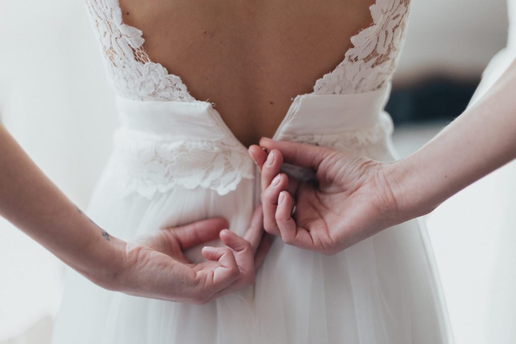 gros plan sur des mains refermant la fermeture d’une robe de mariée en dentelle dans la chambre