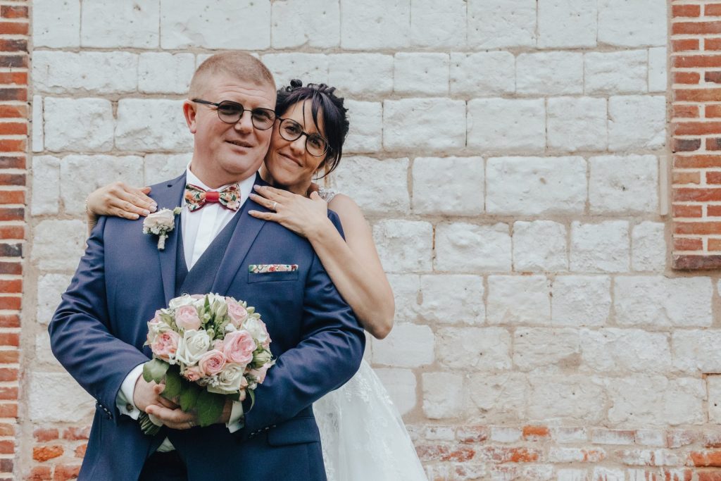mariée enlacée derrière son marié en costume bleu, bouquet de roses devant un mur de briques