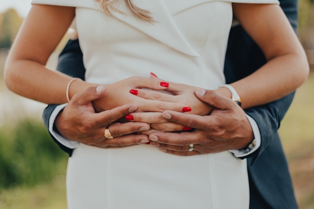 Mains enlacées d’un couple de mariés autour de la taille de la mariée, alliances et vernis rouge bien visibles.