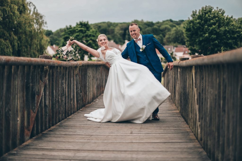 mariés s’amusant sur un pont en bois, la mariée penchée en arrière dans sa robe, le marié la tenant par la main