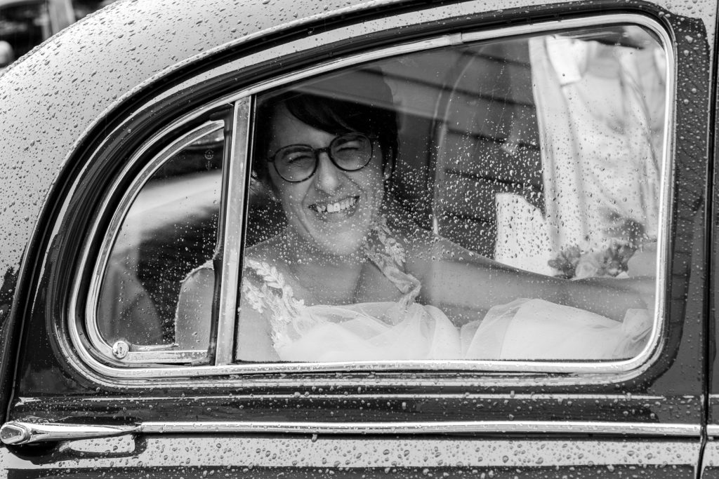 Mariée souriante derrière la vitre mouillée d’une voiture ancienne le jour de son mariage