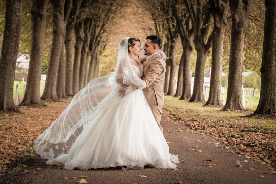 Mariés s’enlaçant dans une allée bordée d’arbres, la longue traîne de la robe balaie le sol couvert de feuilles d’automne.