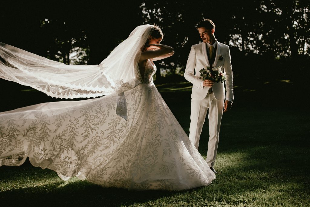 mariée faisant voler son voile en dentelle face à son marié en costume clair – photographie de mariage au coucher du soleil