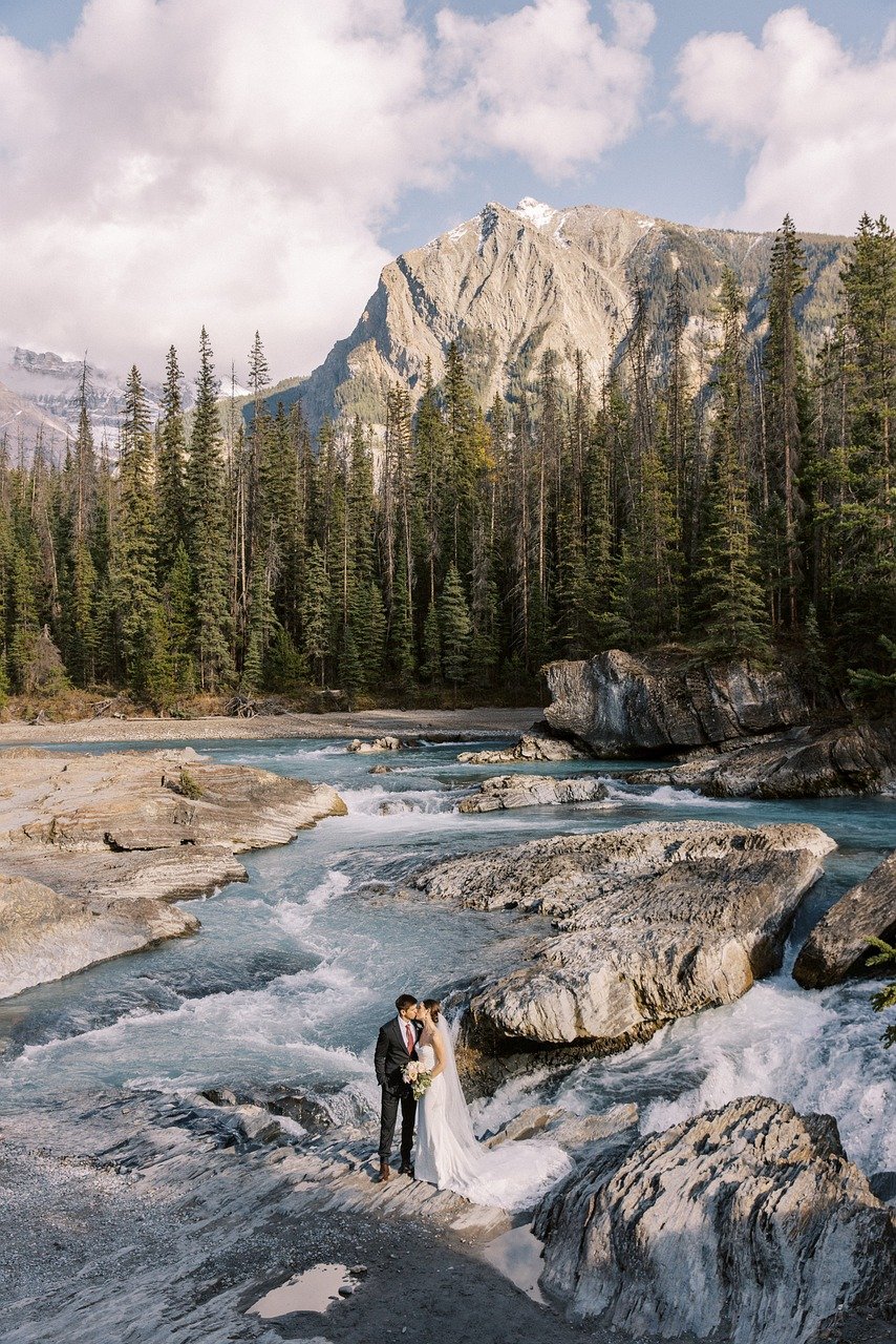 Couple en habits de marié au milieu d'une rivière avec la montagne en arrière plan et des grands sapins.