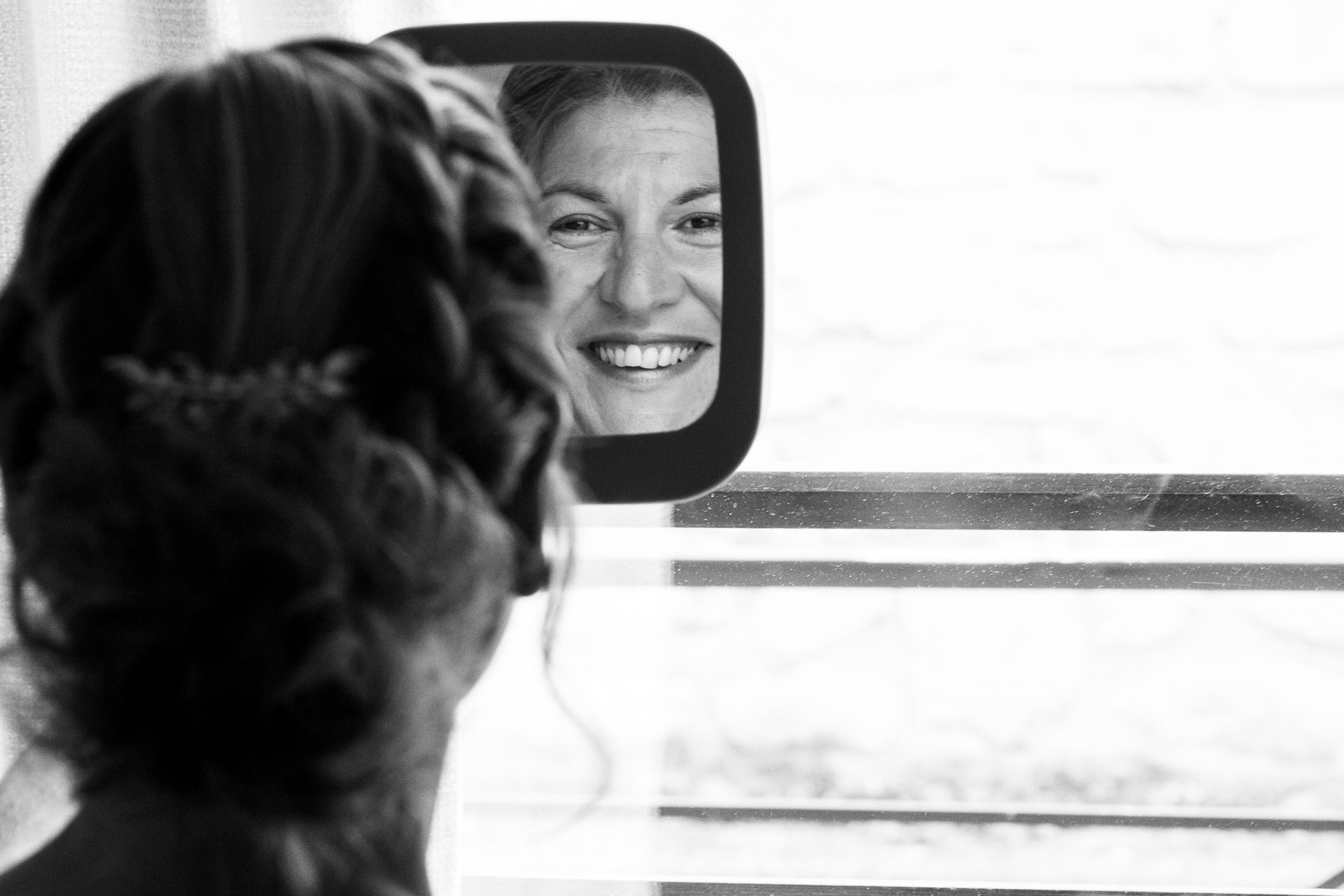Mariée vue de dos, son sourire apparaît dans un petit miroir devant elle, photo de préparation en noir et blanc.