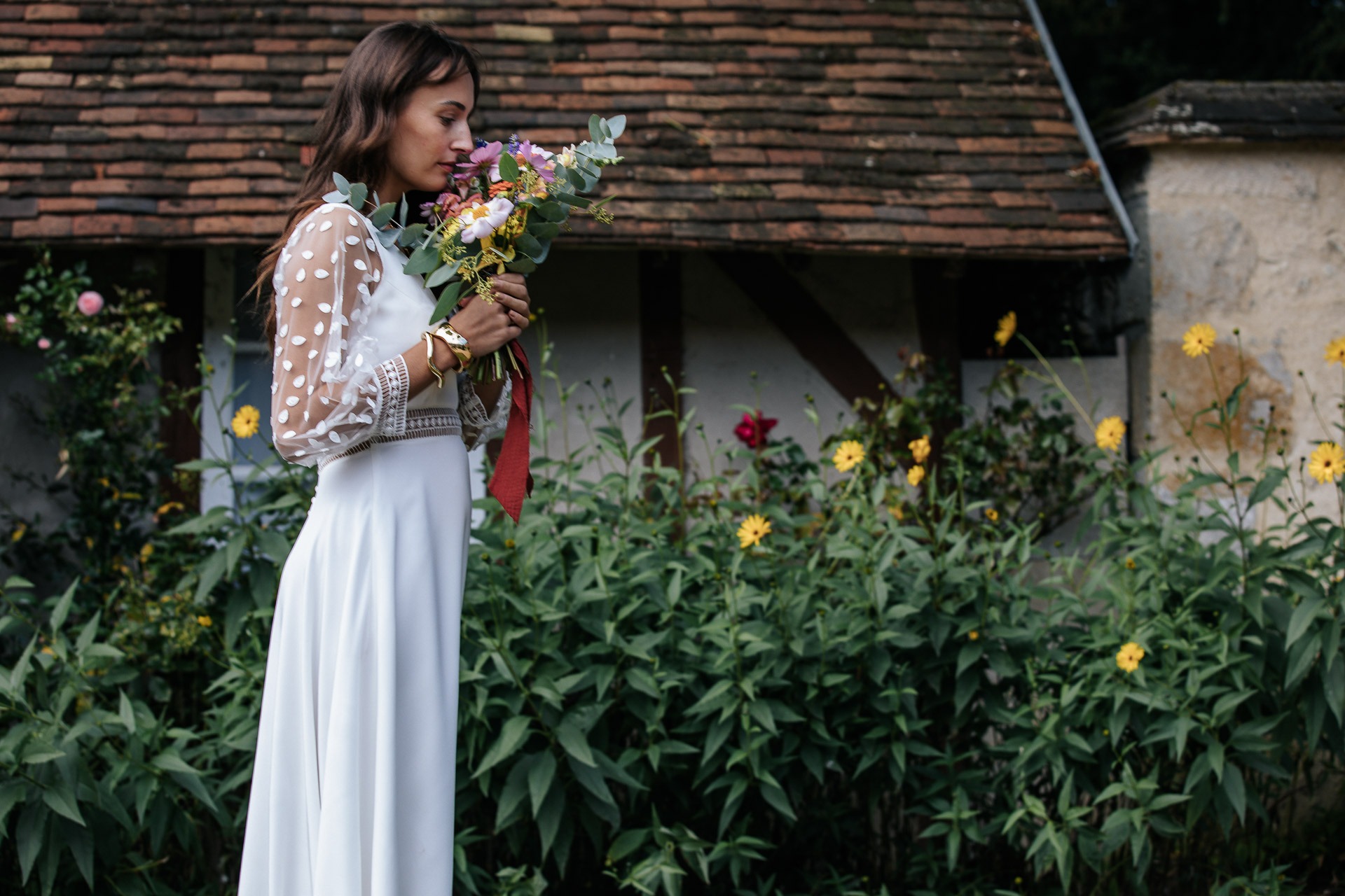 Mariée en robe blanche dans un jardin fleuri, tenant son bouquet et marchant devant une maison de campagne.