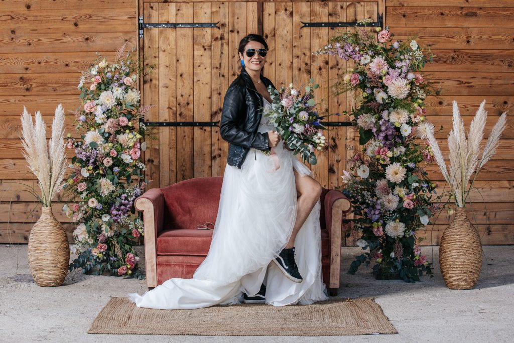 Mariée moderne en veste en cuir et baskets, debout devant un canapé rouge entouré de grandes compositions florales.