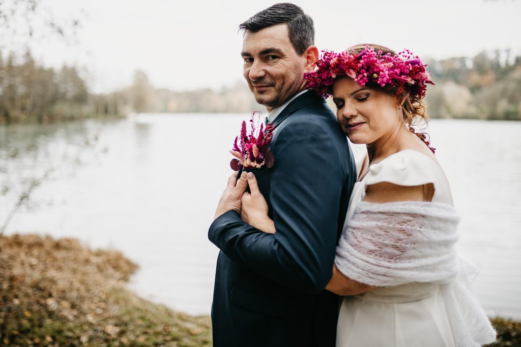 Couple de mariés enlacés au bord d’un lac, la mariée portant une couronne de fleurs fuchsia