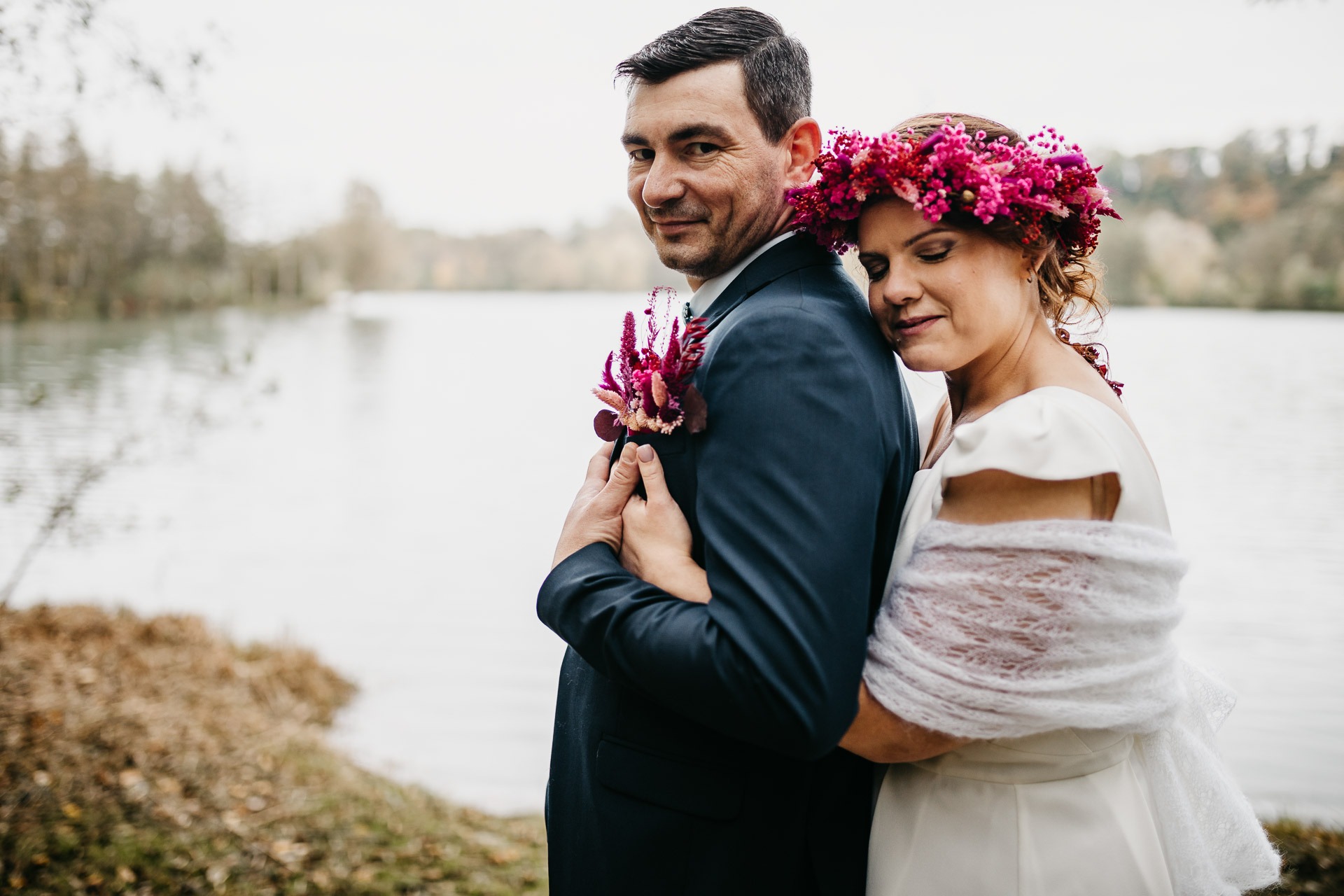 Couple de mariés enlacés au bord d’un lac, la mariée portant une couronne de fleurs fuchsia