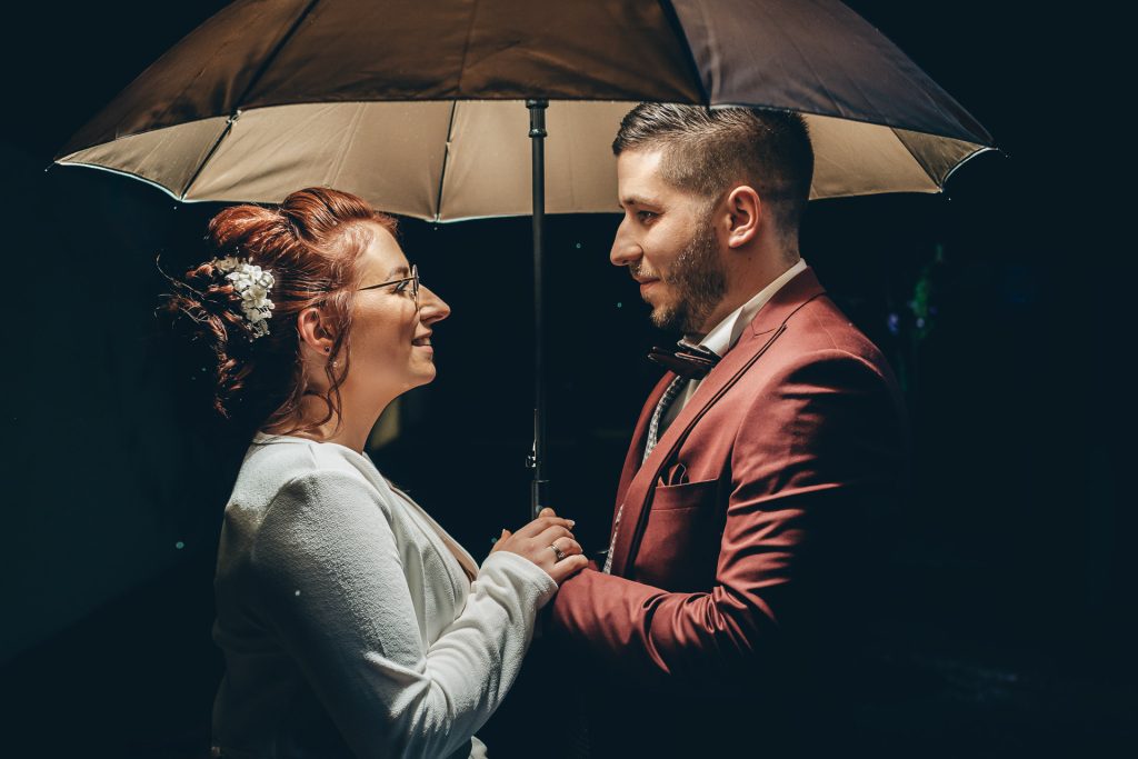Couple de mariés sous un parapluie de nuit, se faisant face et se tenant les mains dans une lumière chaleureuse.