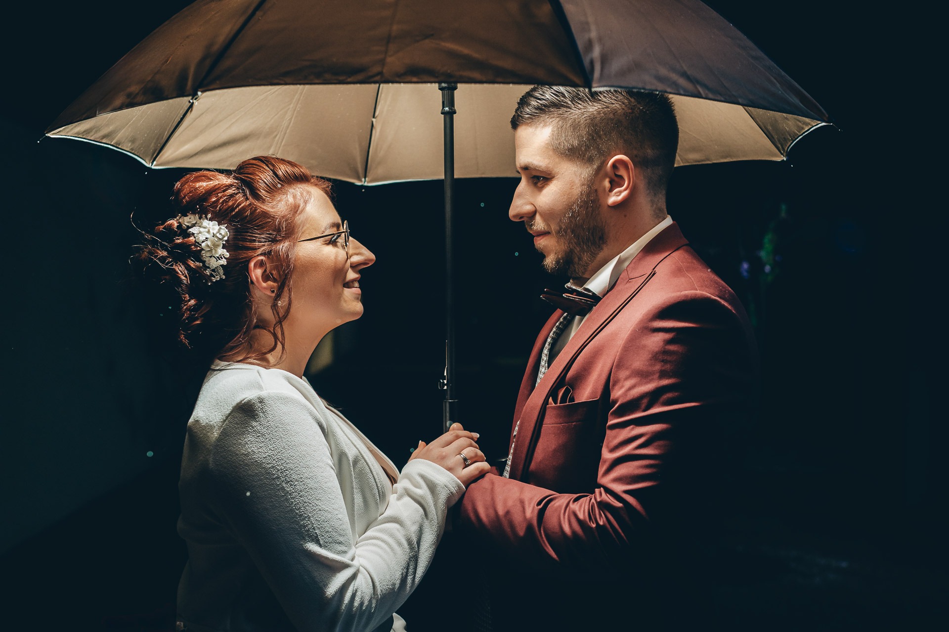 Couple de mariés sous un parapluie de nuit, se faisant face et se tenant les mains dans une lumière chaleureuse.