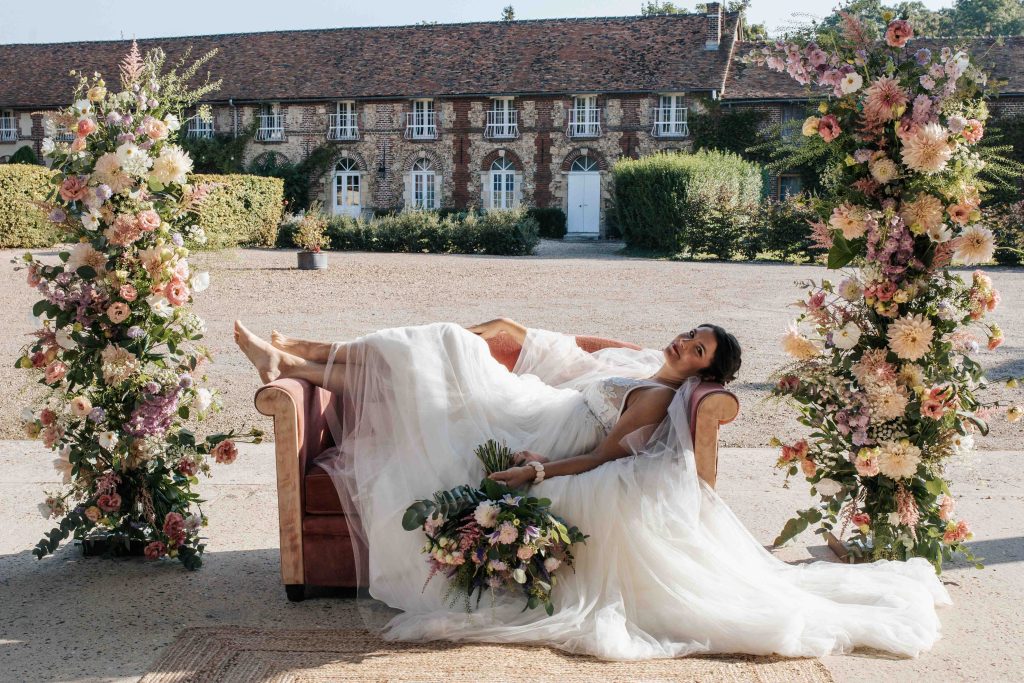 Mariée allongée en robe fluide sur un canapé, entourée de grandes compositions florales dans la cour d’un domaine