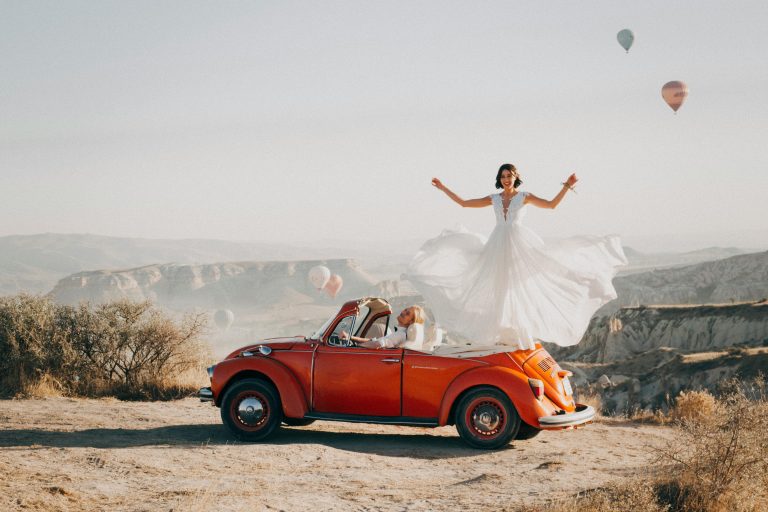 Les mariés célèbrent leur mariage au sommet d'une colline dans une voiture d'époque rouge décapotable, en arrière plan une vue sur les montagnes.