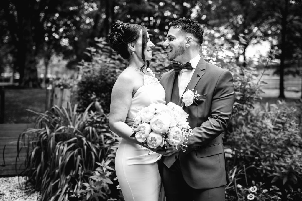 couple de jeunes mariés souriants en noir et blanc, bouquet de roses en premier plan dans un jardin